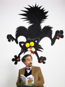 Author Nick Bruel, in a jacket and bowtie, poses with a book underneath a cartoon of his character Bad Kitty.